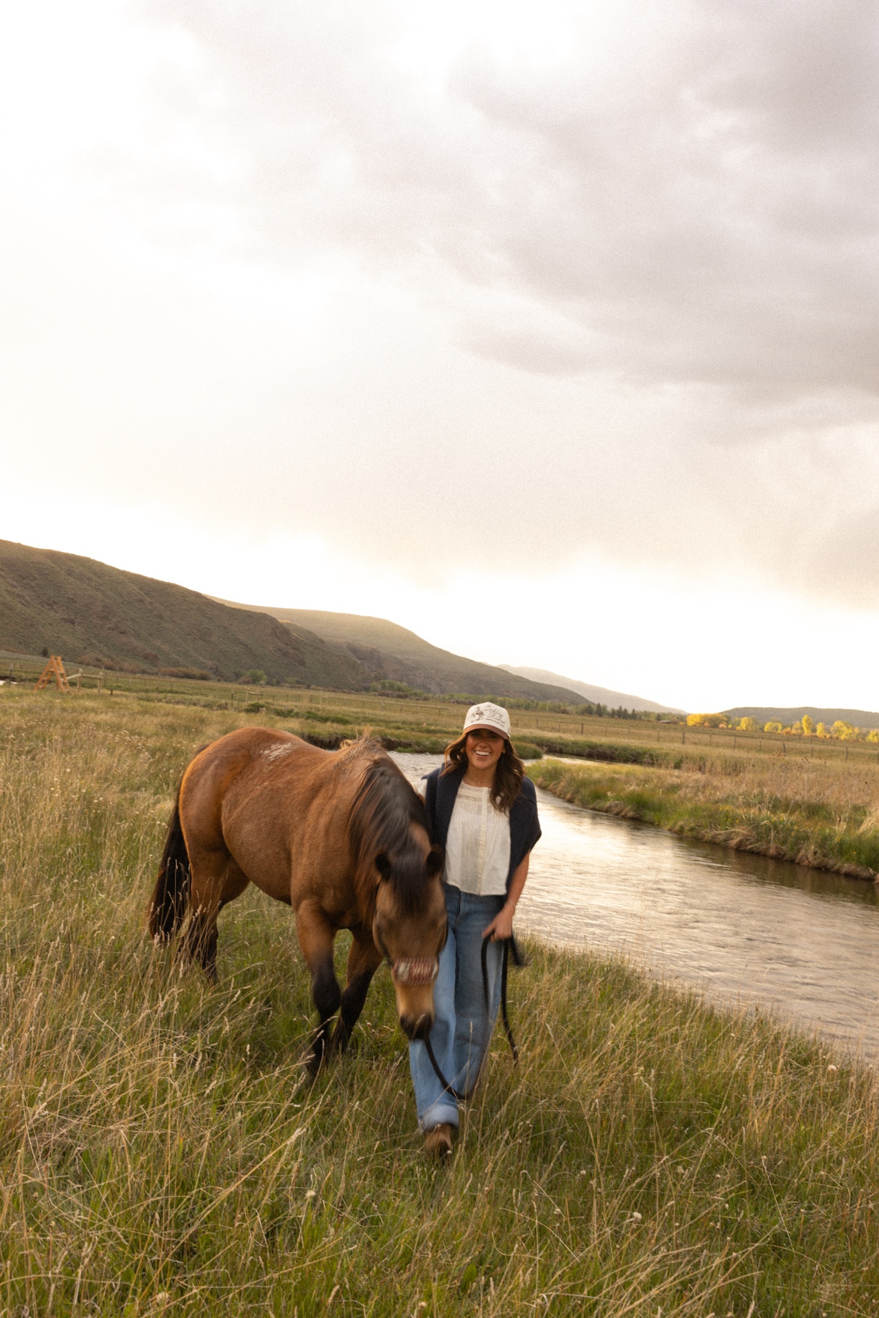 Woman walking a horse along a creek wearing a custom ranch trucker hat at golden hour