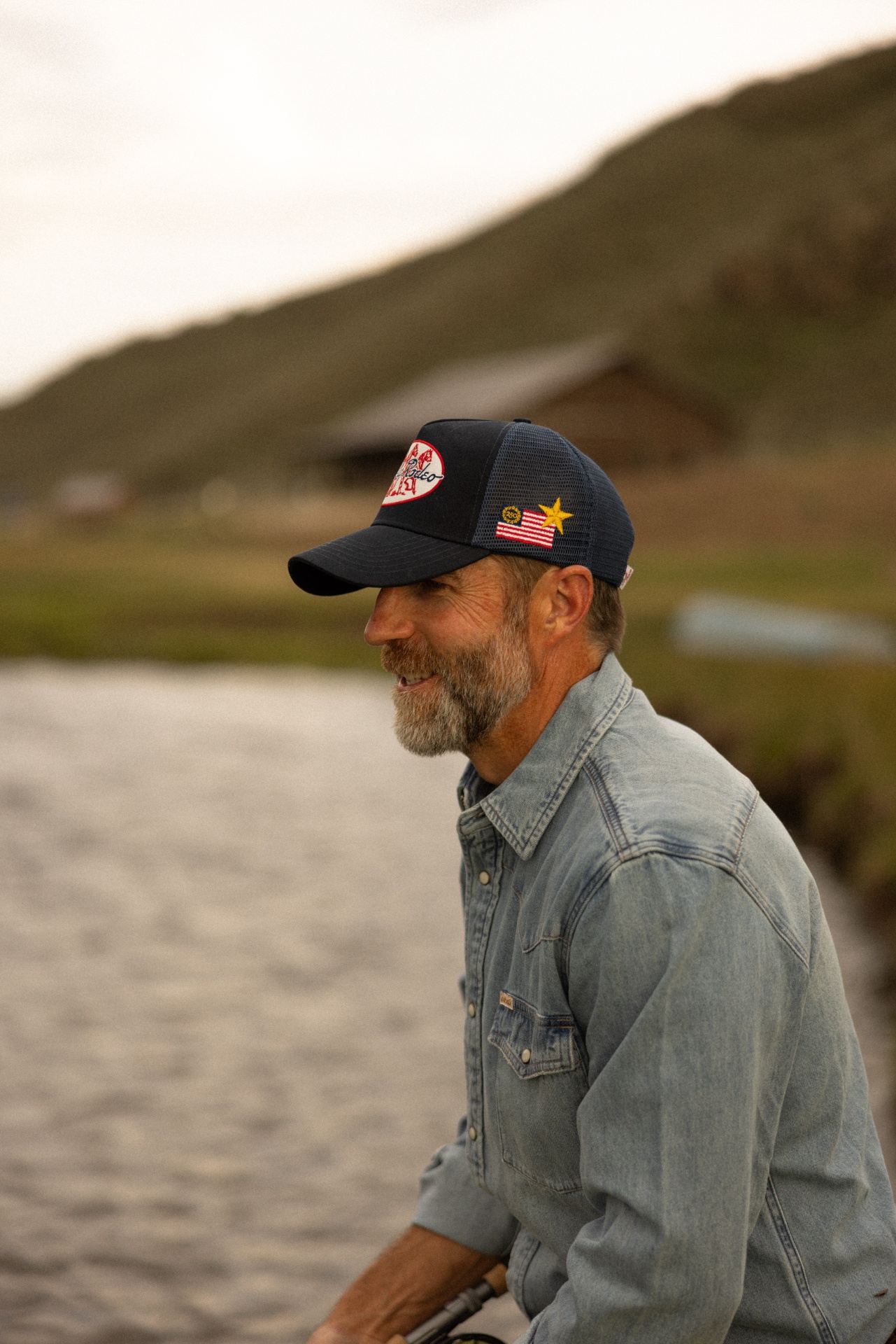 Bearded rancher in denim shirt wearing a navy Oakley Rodeo trucker hat by the creek