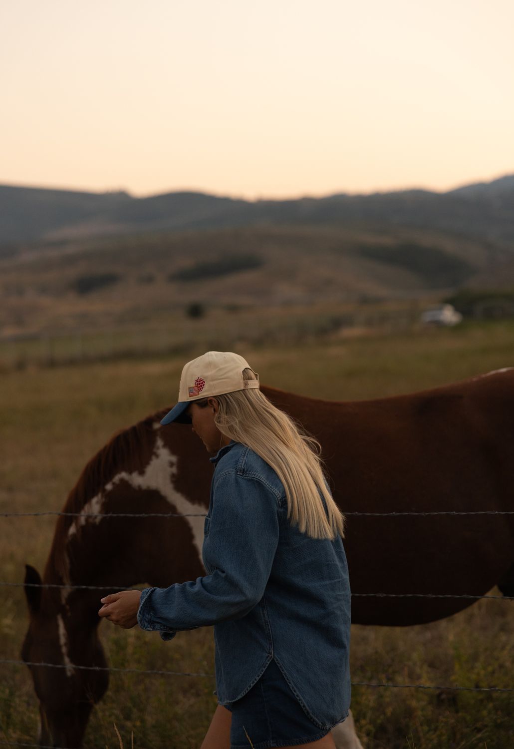 Woman in a rodeo trucker hat leading a paint horse through the pasture at dusk