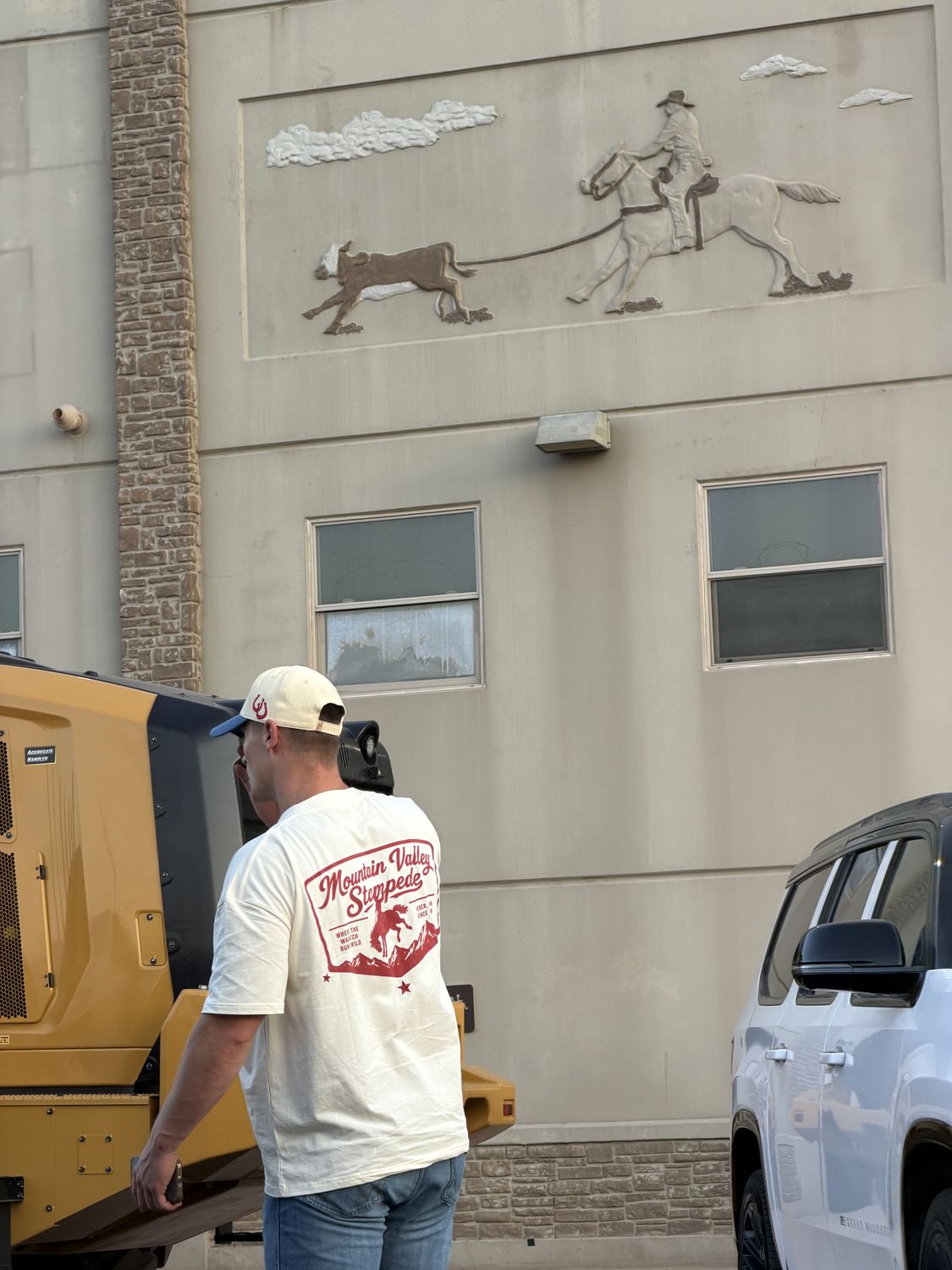 Man in a cream Mountain Valley Stampede tee and matching cap in front of a western roping mural