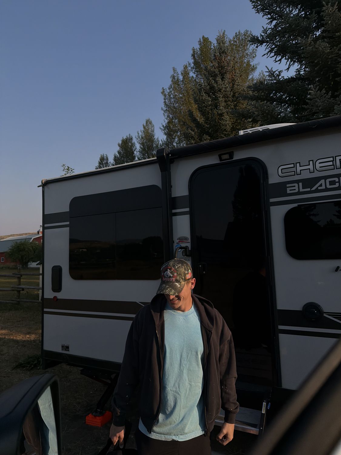 Man in a camo rodeo trucker hat stepping out of the camper at sunset