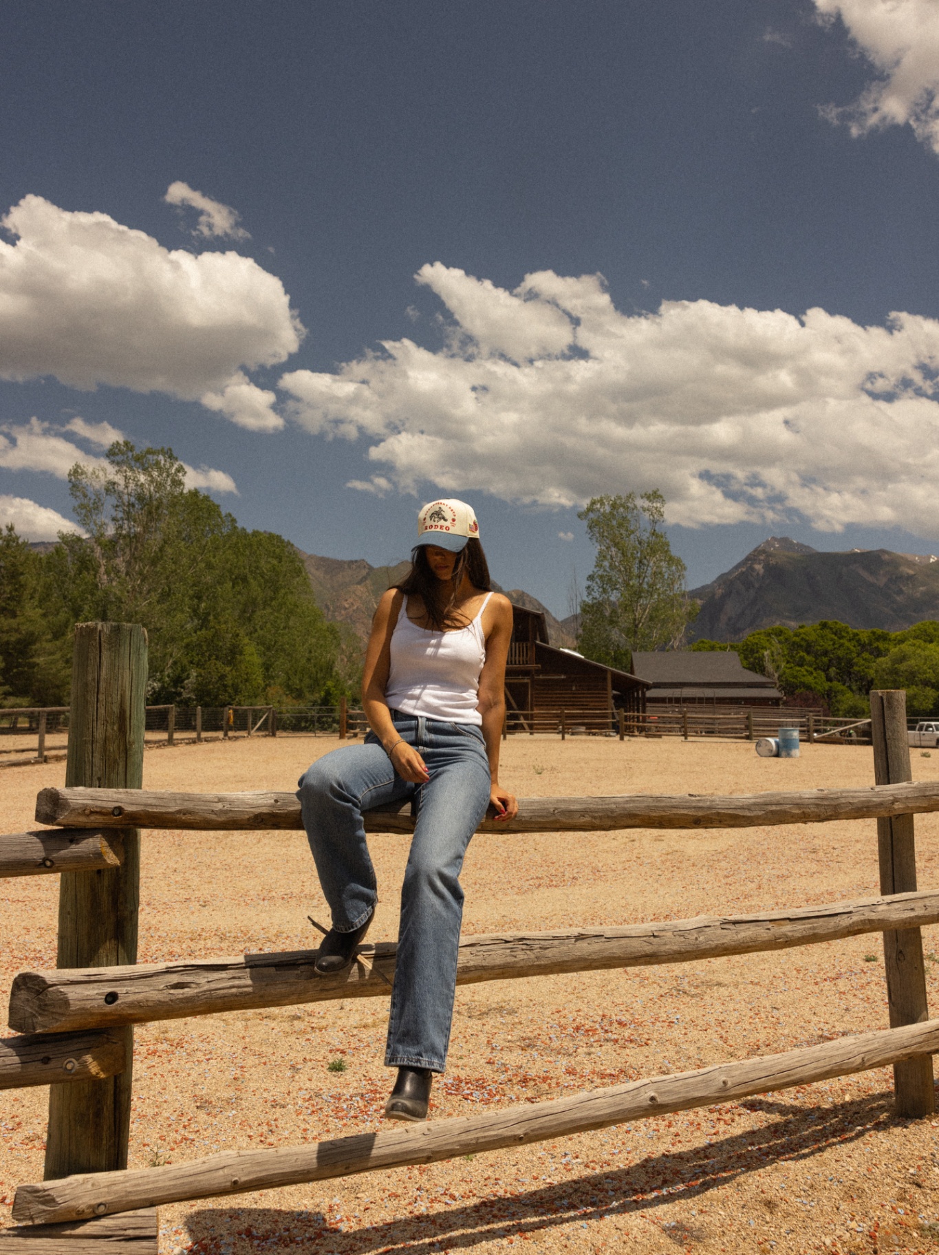 Model on a fence rail in a Strawberry Days Rodeo cap with mountains and barn behind