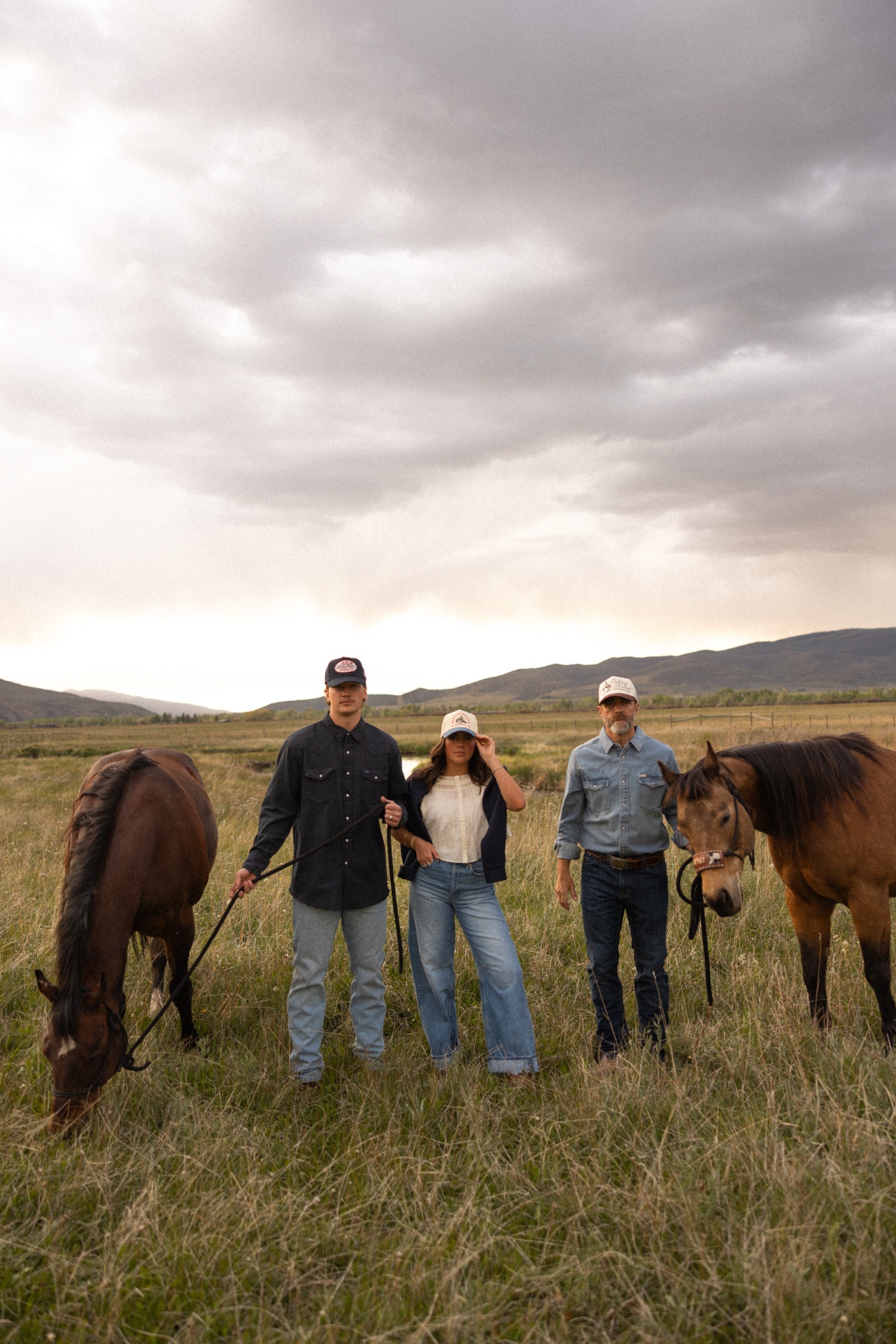 Three models with horses wearing custom Bolt Ranch trucker hats and western shirts under dramatic sky