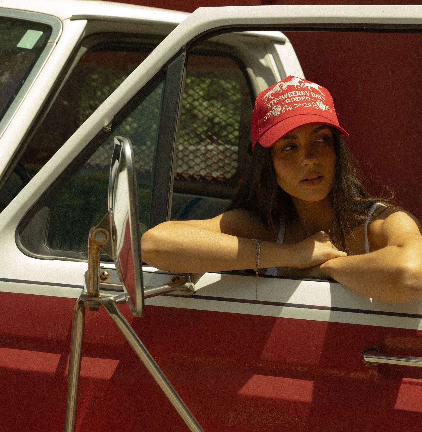 Model in a red Strawberry Days Rodeo trucker hat leaning out the window of a vintage red-and-white truck