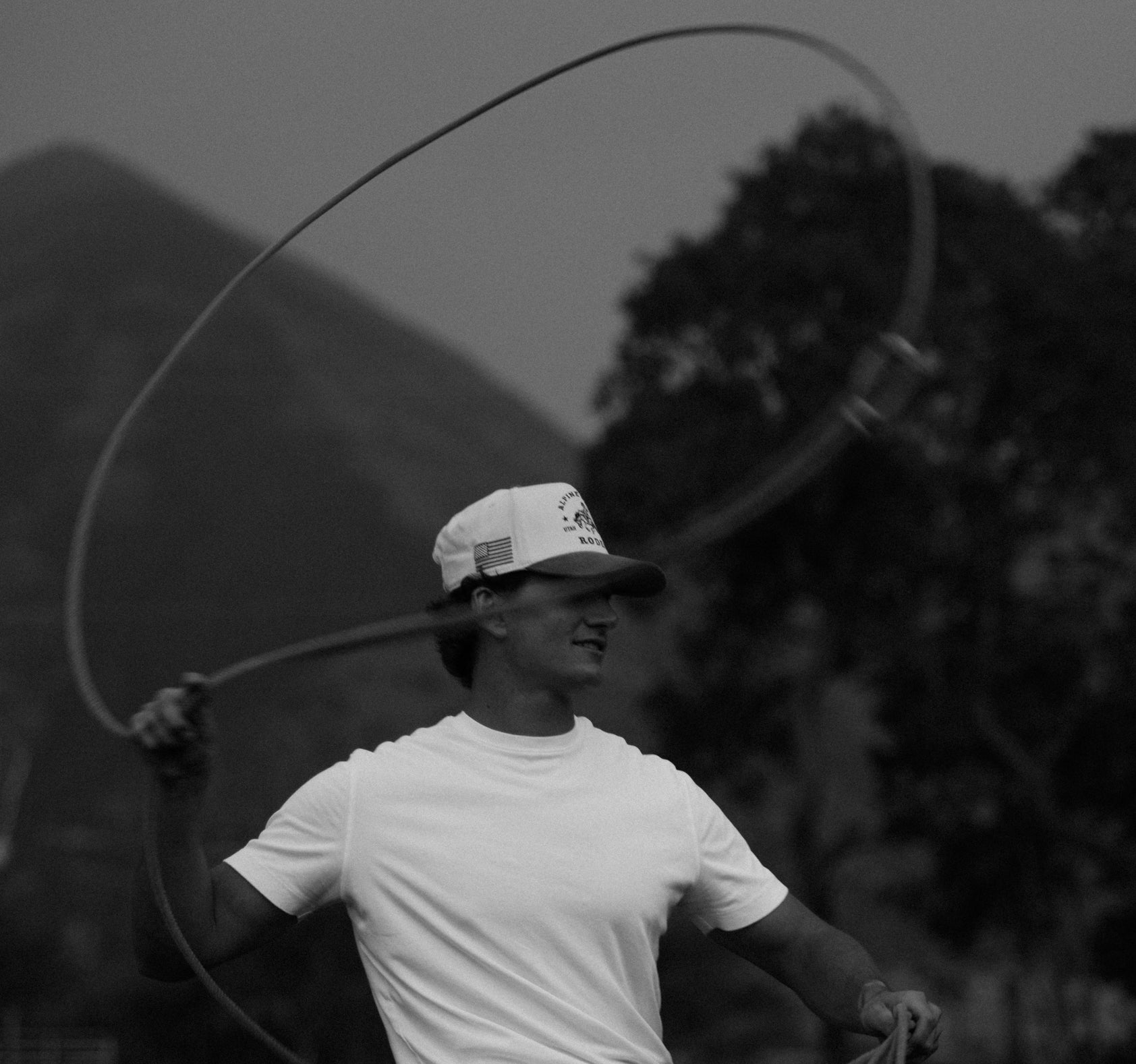 Black and white portrait of a roper mid-swing wearing a custom rodeo hat with mountain backdrop