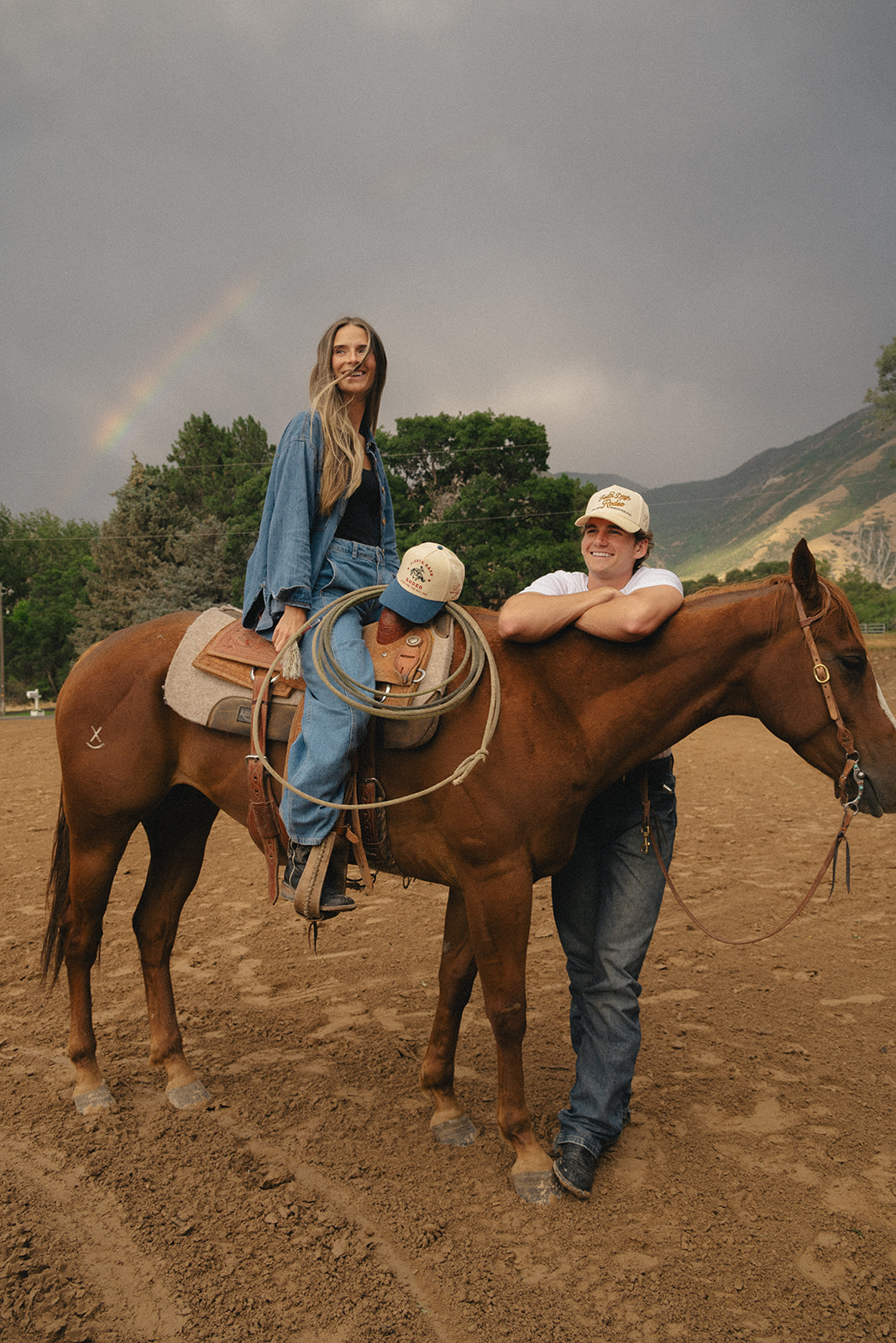 Couple with horse and custom rodeo trucker hats under a rainbow after the storm