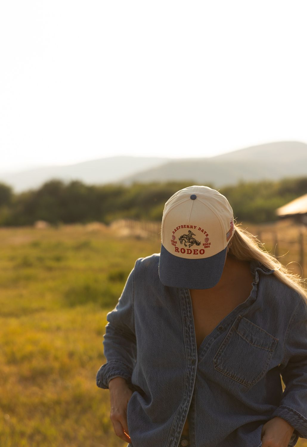 Model in a denim shirt and cream rodeo trucker hat standing in a field at golden hour