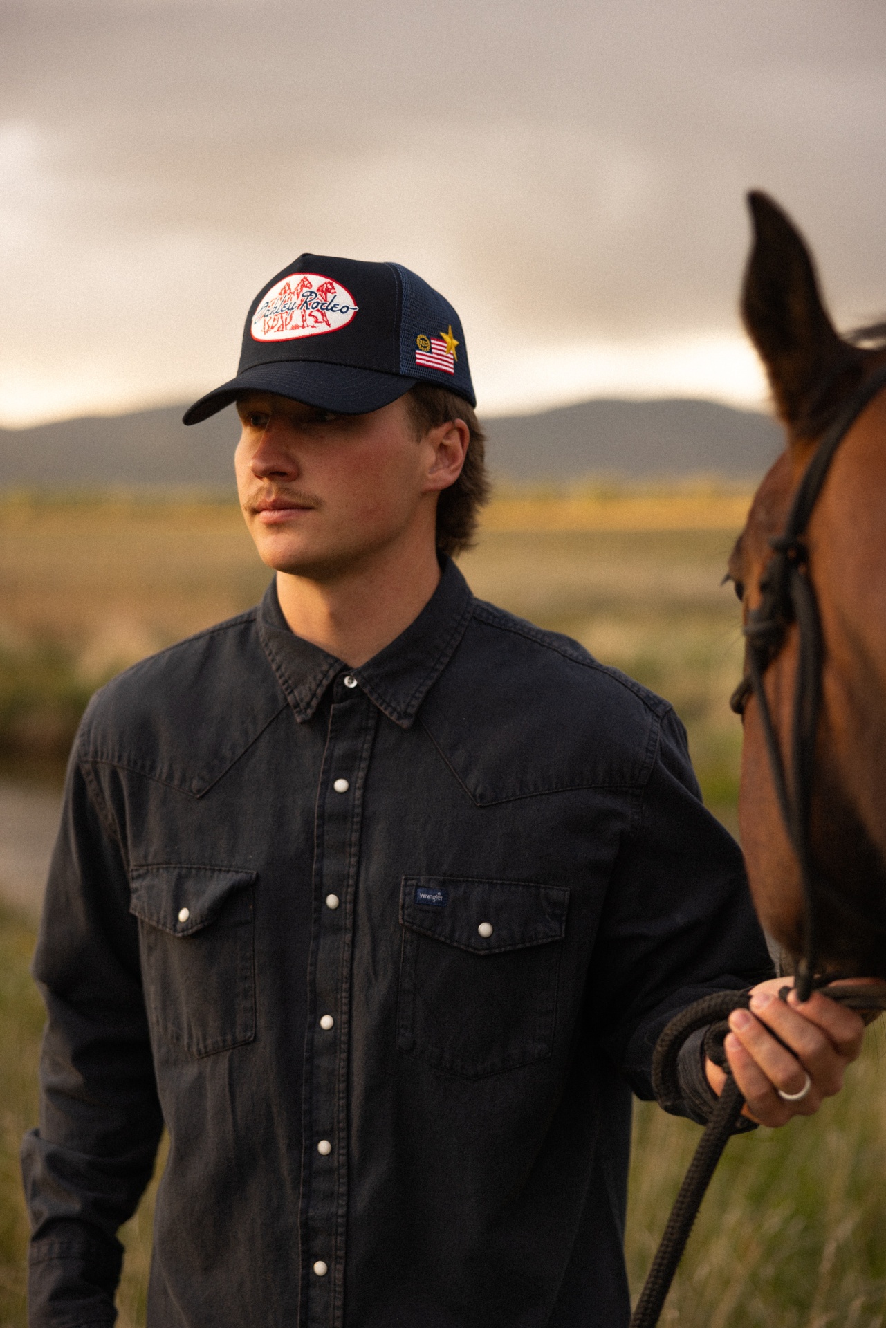 Young man in a dark western shirt and trucker hat holding a horse lead